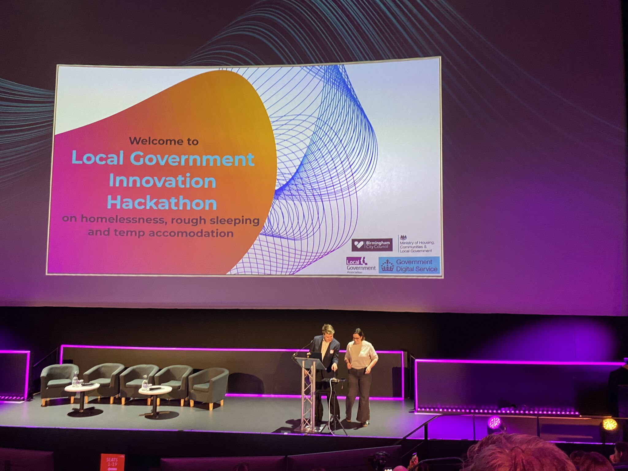 A man and a woman standing at a lectern. The screen behind them reads Welcome to Local Government Innovation Hackathon on homelessness, rough sleeping and temp accommodation